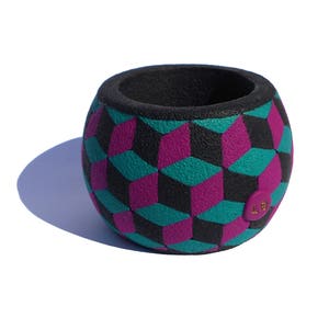 K&ouml;nnte beinhalten: Ein kugelf&ouml;rmiger Beh&auml;lter mit strukturierter Oberfl&auml;che und einem sich wiederholenden geometrischen W&uuml;rfelmuster in Schwarz, T&uuml;rkis und Magenta. Das Innere ist schwarz und ein kleines rundes magentafarbenes Etikett mit goldenen LS-Buchstaben ist seitlich angebracht.