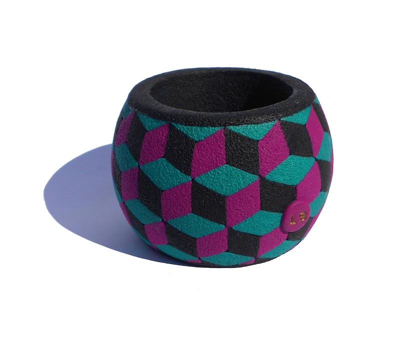 K&ouml;nnte beinhalten: Ein kugelf&ouml;rmiger Beh&auml;lter mit strukturierter Oberfl&auml;che und einem sich wiederholenden geometrischen W&uuml;rfelmuster in Schwarz, T&uuml;rkis und Magenta. Das Innere ist schwarz und ein kleines rundes magentafarbenes Etikett mit goldenen LS-Buchstaben ist seitlich angebracht.