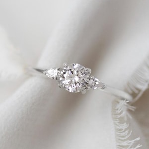 May include: A silver ring with a large, round, clear gemstone in the center, flanked by two smaller, pear-shaped stones. The ring is set against a white, textured fabric with a frayed edge.