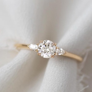 May include: A gold ring with a central round diamond and two smaller side stones. The ring is set on a white fabric background, creating a soft and elegant aesthetic. The ring's design is simple yet sophisticated, suitable for engagement or everyday wear.
