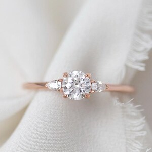 May include: A rose gold engagement ring with a central round diamond and two smaller side stones. The ring is set on a white, textured fabric, creating a soft, romantic aesthetic. The ring's band is delicate and thin.