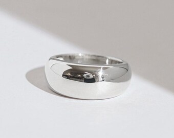 May include: A polished, silver-colored ring with a rounded, smooth design. The ring sits on a white surface, with a shadow cast across the background. The ring's surface reflects the light.