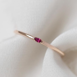 May include: A delicate gold ring with a small, oval-shaped ruby gemstone. The ring is set against a soft, white fabric background, creating a minimalist and elegant aesthetic. The ruby is a vibrant red color.