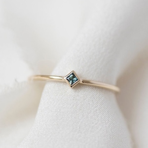 Puede incluir: Un delicado anillo de oro con una pequeña piedra preciosa azul de forma cuadrada.