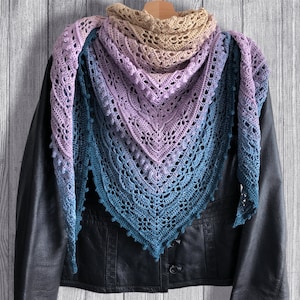 May include: A crocheted shawl with a gradient of colors from beige to purple to blue. The shawl is draped over a black leather jacket.