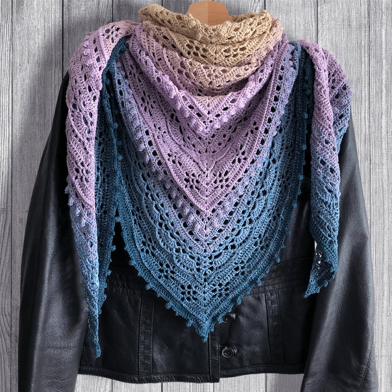 May include: A crocheted shawl with a gradient of colors from beige to purple to blue. The shawl is draped over a black leather jacket.