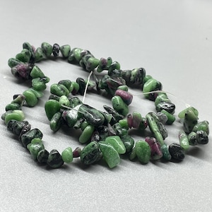 May include: Strand of natural ruby zoisite chip beads. The beads are a mix of green, black, and pinkish-red colors. The beads are strung on a white string and are suitable for jewelry making.