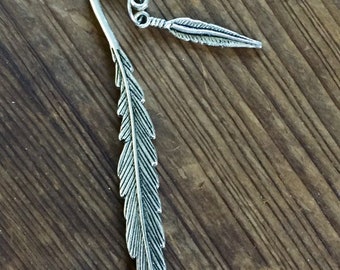May include: A silver-colored metal bookmark in the shape of a feather. The bookmark has a curved top with decorative swirls and a smaller feather charm attached. The main feather design is detailed with etched lines, creating a realistic appearance.