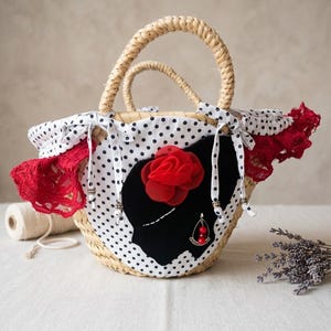 Könnte beinhalten: Eine Stroh-Tragetasche mit geflochtenen Griffen, verziert mit einer schwarzen Silhouette, einer roten Stoffrose und einem roten Perlenohrring. Die Tasche hat einen weißen Stoff mit schwarzen Punkten und roten Spitzensaum.
