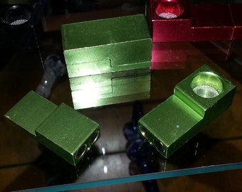 May include: Several green metal pipes are displayed on a glass surface. The pipes have a rectangular design with a circular opening and a mesh screen. One pipe is red. The pipes are arranged in different orientations.