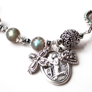 May include: A silver-toned bracelet featuring a mix of beads and charms. The bracelet includes round, gray-green beads, decorative silver beads, and several charms, including a cross, a heart, and a religious medallion. The bracelet has a decorative clasp.