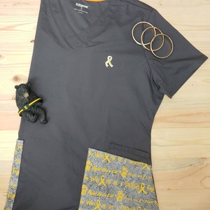 May include: Gray scrub top with a yellow ribbon embroidered on the chest. The top has a V-neck and short sleeves. The pockets have a gray and yellow print with the text "You are a Warrior, Fight!" repeated.