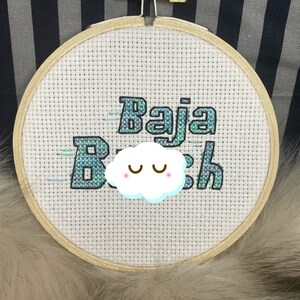 May include: A white embroidery hoop with the text "Baja Beach" in blue and green with a white cloud with eyes above the text.
