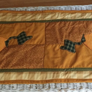 May include: A patchwork table runner with a fall theme. The runner is made of various shades of brown, orange, and green fabrics. It features a checkered pattern and a green border.