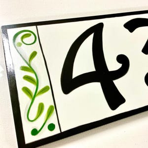 May include: White ceramic house number sign with black cursive numbers 43 and green floral designs on the sides.