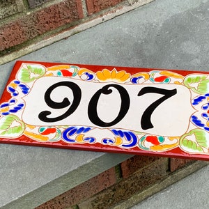 May include: A rectangular ceramic house number plaque featuring the digits 907 in black. The plaque has a cream-colored center surrounded by a decorative border with red, orange, blue, and green floral and leaf patterns on a deep red background.
