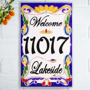 May include: A white ceramic welcome sign with a blue background and colorful floral accents. The sign features the text "Welcome" and the number "11017" in black. The text "Lakeside" is also in black.