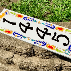 May include: Rectangular white ceramic address plaque with the number 1455 in black. The border features a colorful design with orange, blue, green, and yellow accents. The plaque is placed on a stone surface.
