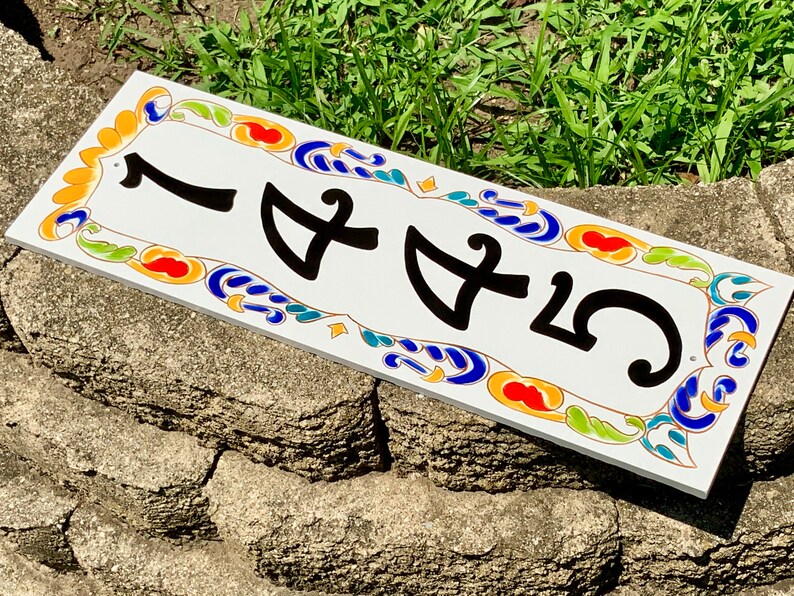 May include: Rectangular white ceramic address plaque with the number 1455 in black. The border features a colorful design with orange, blue, green, and yellow accents. The plaque is placed on a stone surface.
