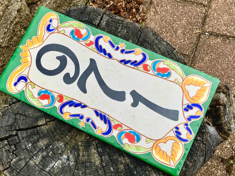 May include: Rectangular ceramic house number plaque with the number 27 in black on a cream background. The plaque has a green border and is decorated with colorful floral and scroll designs. The plaque is resting on a weathered tree stump.