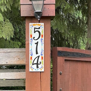 May include: A rectangular address plaque with the numbers "514" in black. The plaque is white with colorful floral designs and is mounted on a wooden post. A black lantern hangs above the plaque.