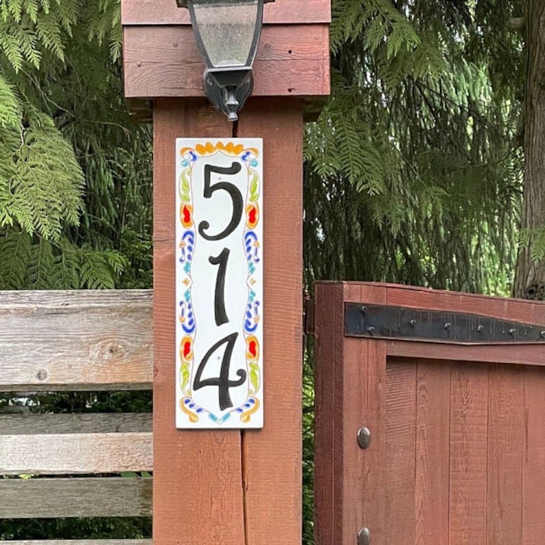 May include: A rectangular address plaque with the numbers "514" in black. The plaque is white with colorful floral designs and is mounted on a wooden post. A black lantern hangs above the plaque.