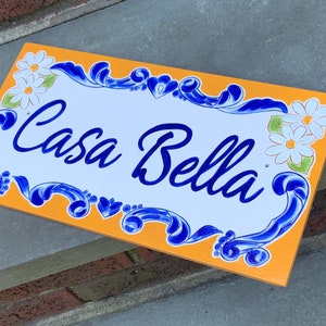 May include: A yellow and white ceramic sign with blue floral accents and the text "Casa Bella" in blue cursive lettering.