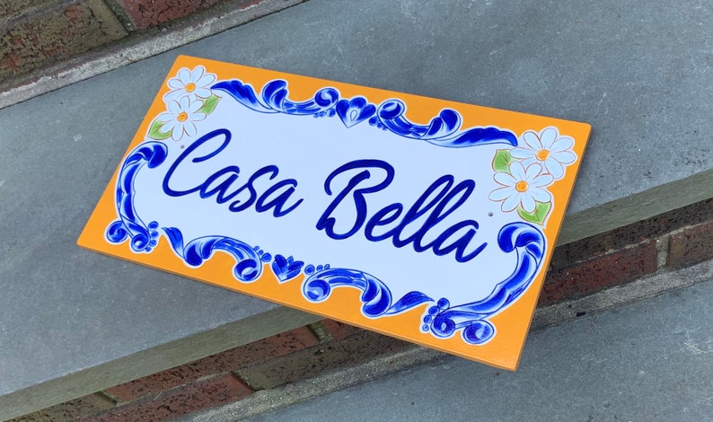 May include: A yellow and white ceramic sign with blue floral accents and the text "Casa Bella" in blue cursive lettering.