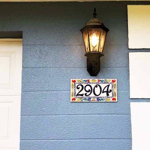 May include: A blue house with a white door and a black light fixture. A ceramic house number plaque with colorful floral designs reads "2904".