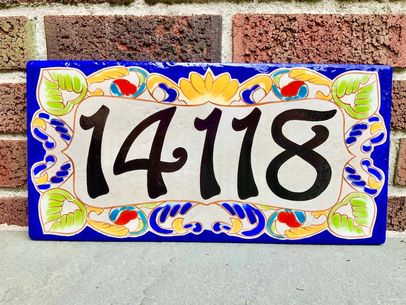 May include: A rectangular ceramic house number plaque featuring the black digits 14118 centered on a cream background. The border is decorated with a vibrant pattern of green leaves, yellow floral motifs, and red accents against a deep blue frame.