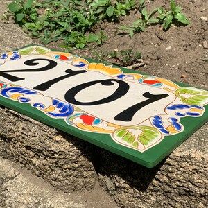 May include: A rectangular house number plaque featuring the black digits 2101 centered on a cream-colored background. The border is decorated with a colorful floral pattern in shades of green, blue, orange, and yellow, set against a dark green frame.