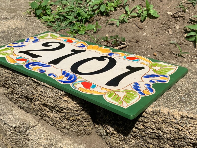 May include: A rectangular house number plaque featuring the black digits 2101 centered on a cream-colored background. The border is decorated with a colorful floral pattern in shades of green, blue, orange, and yellow, set against a dark green frame.