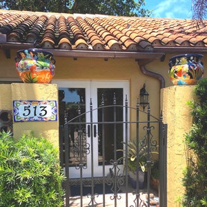 May include: A house entrance featuring a yellow stucco wall with a ceramic tile address plaque displaying the number 513. Two colorful hand-painted ceramic planters with floral motifs sit atop the pillars flanking a black wrought iron gate.