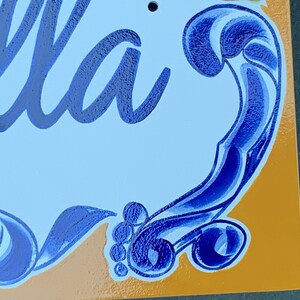 May include: A blue and white ceramic sign with the word "Bella" written in cursive. The sign has a decorative blue swirl design around the edges.