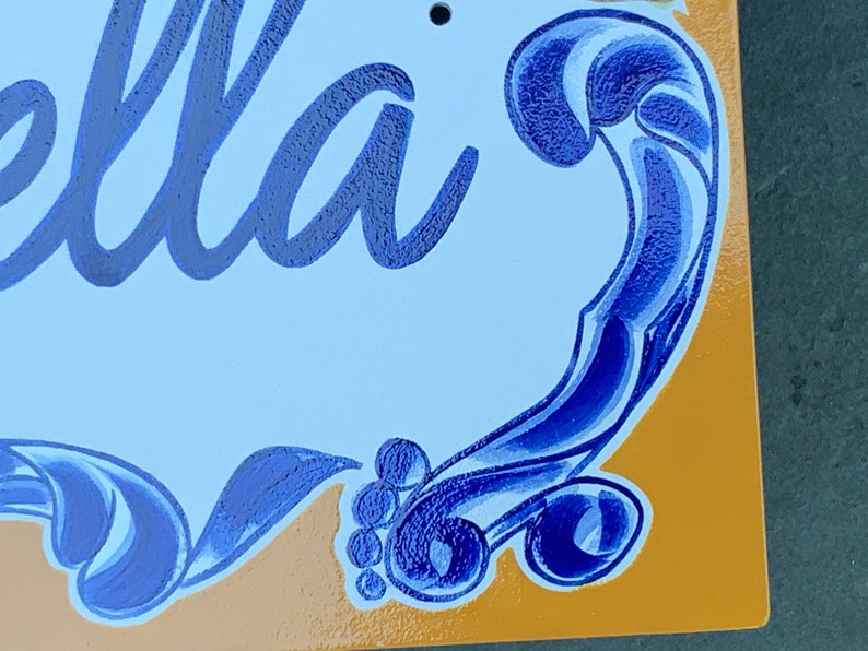 May include: A blue and white ceramic sign with the word "Bella" written in cursive. The sign has a decorative blue swirl design around the edges.