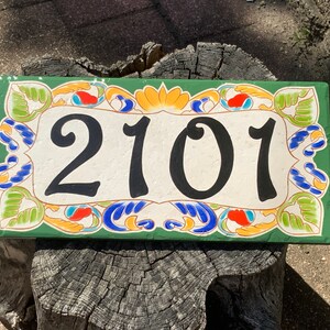 May include: A rectangular ceramic house number plaque featuring the black digits 2101 centered on a cream background. The plaque is framed by a decorative border of green leaves, orange petals, and blue swirling accents on a dark green base.