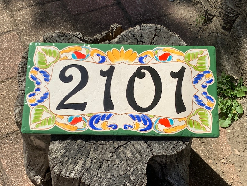 May include: A rectangular ceramic house number plaque featuring the black digits 2101 centered on a cream background. The plaque is framed by a decorative border of green leaves, orange petals, and blue swirling accents on a dark green base.