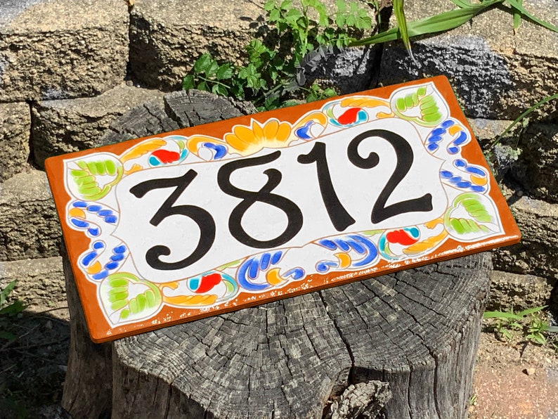 May include: A rectangular ceramic house number plaque featuring the black digits 3812 centered on a white background. The border is decorated with a hand-painted floral pattern in vibrant shades of orange, yellow, blue, and light green.