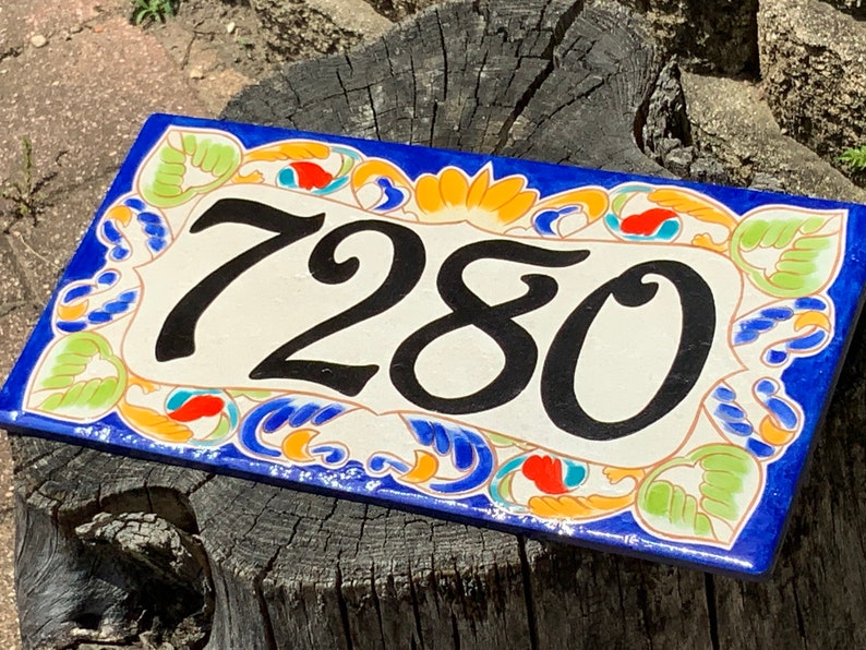 May include: A rectangular ceramic house number plaque featuring the black digits 7280 centered on a cream background. The border is decorated with a hand-painted pattern of blue, orange, and light green floral and leaf motifs on a dark blue base.
