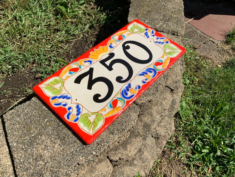 May include: A rectangular ceramic house number plaque featuring the digits 350 in black. The plaque has a cream-colored center and a vibrant red border decorated with hand-painted green leaf motifs and blue and orange scrollwork patterns.