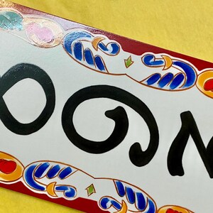 May include: Rectangular ceramic house number plaque with the number 803 in black script. The white plaque has a decorative border with red, blue, orange, and green accents. The background is yellow.