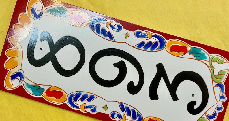 May include: Rectangular ceramic house number plaque with the number 803 in black script. The white plaque has a decorative border with red, blue, orange, and green accents. The background is yellow.