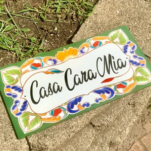 May include: A rectangular sign with a dark green border and a white center featuring the black text Casa Cara Mia. The border is decorated with a colorful pattern of green leaves, orange flowers, and blue swirling accents in a painted style.