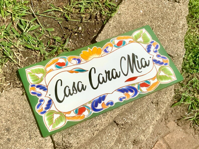 May include: A rectangular sign with a dark green border and a white center featuring the black text Casa Cara Mia. The border is decorated with a colorful pattern of green leaves, orange flowers, and blue swirling accents in a painted style.
