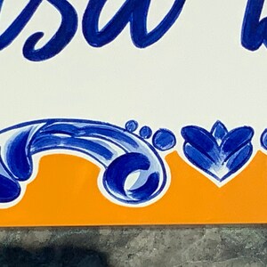 May include: A white tile with blue and orange painted designs. The tile has the word "Casa" written in blue cursive.