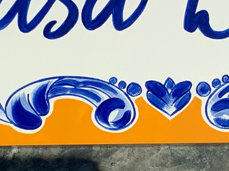 May include: A white tile with blue and orange painted designs. The tile has the word "Casa" written in blue cursive.