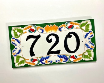 May include: A rectangular house number plaque featuring the black digits 720 centered on a white background. The border is painted in a deep green with decorative floral and leaf motifs in shades of orange, blue, and light green.
