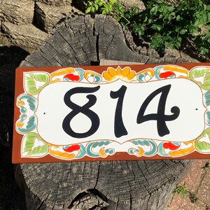 May include: A rectangular house number plaque with a white center featuring the black digits 814. The border is decorated with a colorful floral and leaf pattern in shades of green, orange, and teal against a solid brown background.