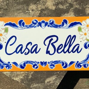 May include: A white ceramic sign with blue and orange accents. The sign reads "Casa Bella" in a decorative script font. The sign is decorated with white daisies.