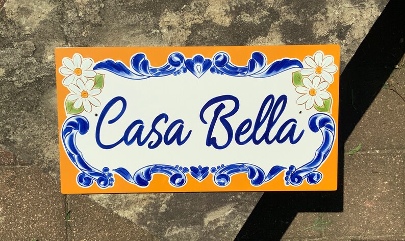 May include: A white ceramic sign with blue and orange accents. The sign reads "Casa Bella" in a decorative script font. The sign is decorated with white daisies.